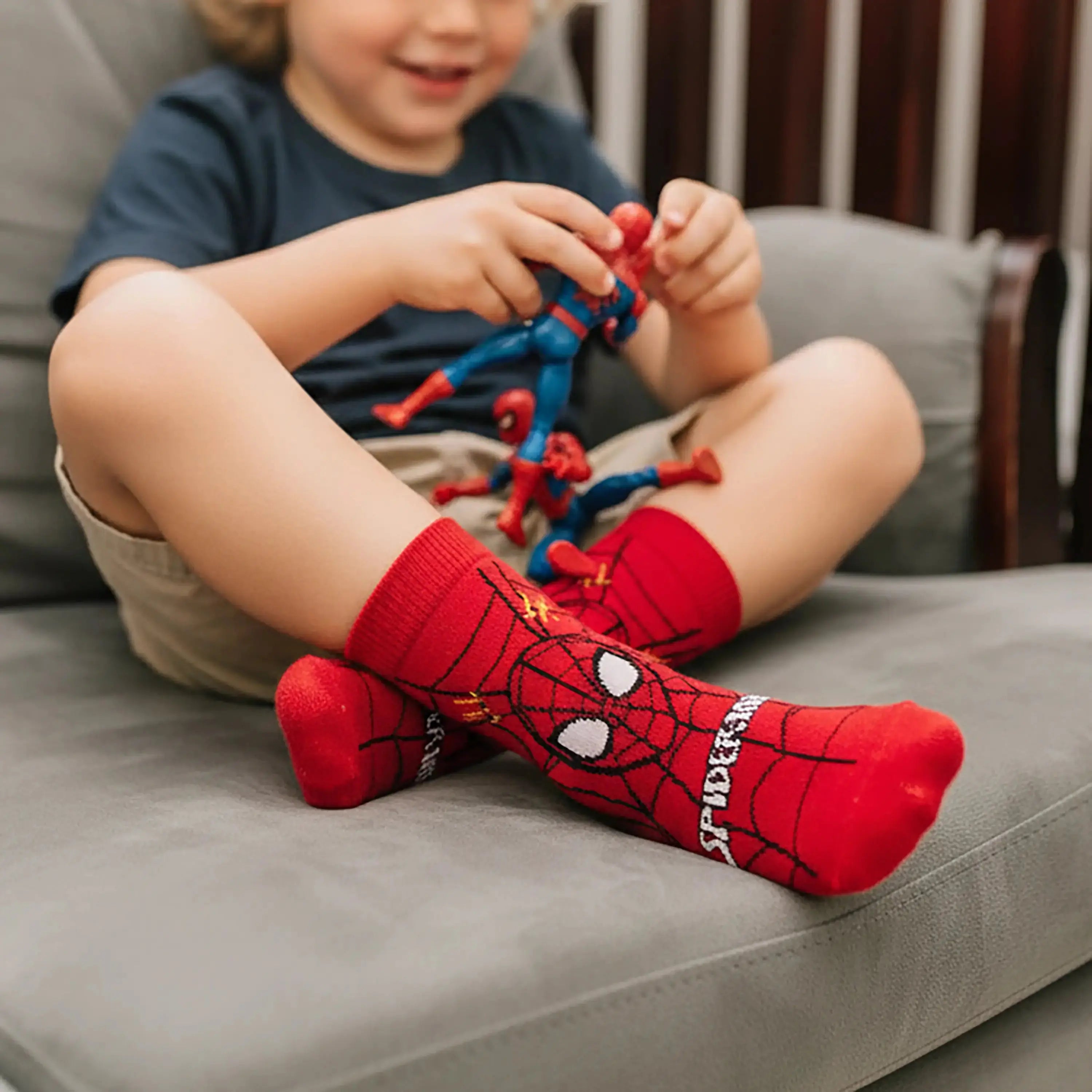 An image of a boy wearing Supersox Marvel Spider-Man Red Printed Crew Socks while playing with a Spider-Man action figure on a sofa