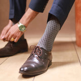 An image of a man wearing black formal socks with white dots and brown leather dress shoes while adjusting his trousers.